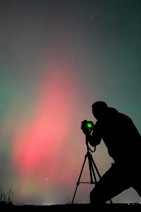 Andy Roe A man is silhouetted taking a picture of the pink and blue Northern Lights