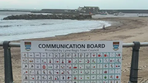 A large board attached to the railings on the beach. It has 144 squares, each containing a commonly used word or number with an illustration. Behind the sign is the sandy beach and sea with the Cobb harbour wall in the background.