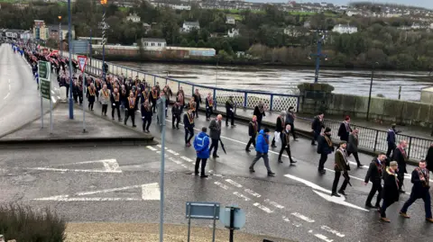 Many men are parading across a bridge. They are wearing sashes and some are carrying umbrellas.