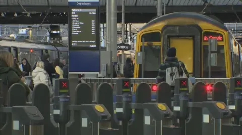 There are two trains in the station. There are security barriers in the foreground and passengers in the background.