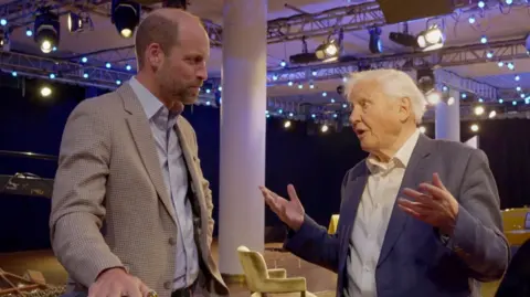 Prince William and David Attenborough have a conversation, standing up in what looks like a television studio with rigged ceiling lights and white pillars in the background. They are standing and William is on the left, a head taller than Sir David and wearing a beige checkered sports jacket and open blue shirt. He looks intently at David, who is wearing a dark blue sports jacket and open white shirt, and has his hands up and open, seeming to emphasise an important point. A look of serious concern is on his face as he looks directly at William, mouth open in mid conversation..