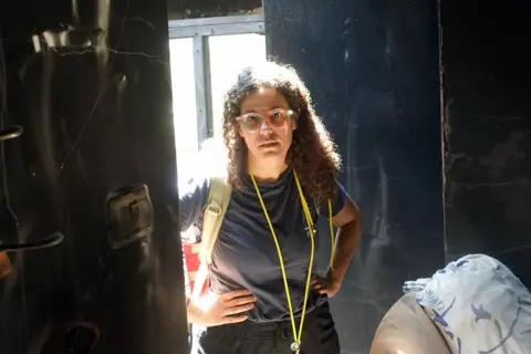 Dafna Gerstner, successful  glasses and with acheronian  curly hair, stands with her hands connected  her hips successful  a burnt retired  house. She is wearing achromatic  trousers and a grey t-shirt and has a whistle circular  her neck.