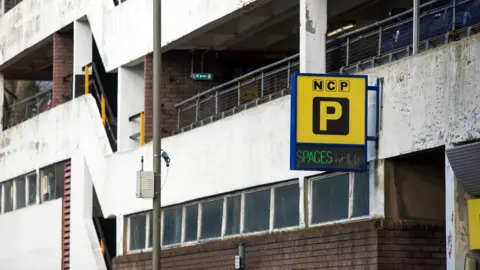 Bloomberg via Getty Images The exterior of a multi-story car park is seen with a sign the has the 'P' logo indicating parking, with the letters NCP above and 'spaces' below, taken in London in 2021.