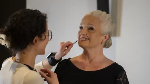 Step Change Studios A woman, Deborah Impiazzi, wearing a black dress. She is getting her makeup done by another woman, left, who is holding a brush up to her lips.
