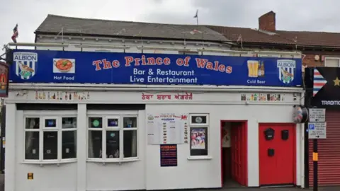 A white building with bay windows and a red door with The Prince of Wales in red writing on a blue sign. There are two bay windows, to the left of the door, with the exterior of the building painted white.