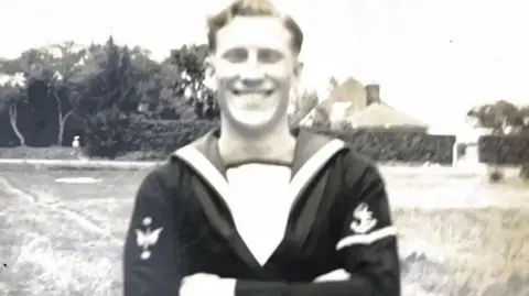 FAMILY PHOTO A black and white photo of a young John Daniels, wearing naval uniform with the characteristic sailor's collar playing out flat towards his shoulders. He is standing in a field with a house in the background. His arms are folded and he is grinning at the camera. 