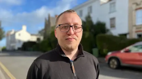 Andy Fletcher is wearing black-framed glasses and a black zip-neck top and standing in a residential street in Exeter.