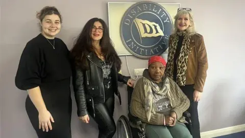 Four women, one of whom is using a wheelchair, are inside an office. They are in front of a gold sign with the charity name on it mounted on the wall.