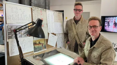 Tom MacDougall/BBC Identical twins Nigel and Klive - wearing matching beige jackets - are pictured at a recreation of their father, Arthur's, animating desk. It is backlit, meaning that the drawing on the desk is lit from underneath. Pinned on a board behind the desk are sketches of animals from Watership Down.