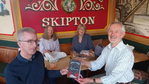 Skipton Esperanto Group Four people sat at a pub table with a big red sign behind them reading The Boat House Skipton 
