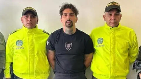 X/@DirectorPolicia Emanuele Gregorini, his hands cuffed behind his back and wearing a black Milan football shirt, is paraded by police in high-vis jackets. 