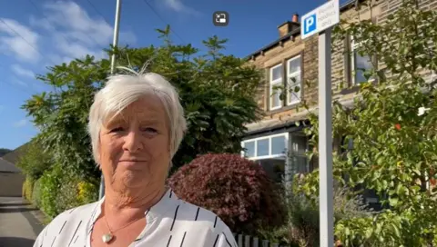 Aisha Iqbal/BBC Christine Fox stands in front of a permit holders-only sign. She has short white hair and is wearing a white blouse with black stripes.