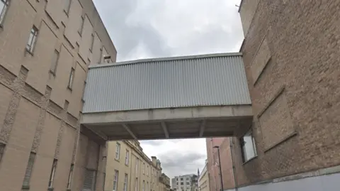 Google A concrete bridge links two sand-coloured buildings