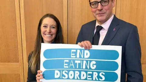 Hope wearing a black dress with long dark hair standing next to Scott, wearing a dark-coloured suit and tie, standing in front of wooden doors and holding a white and blue sign that says End Eating Disorders.