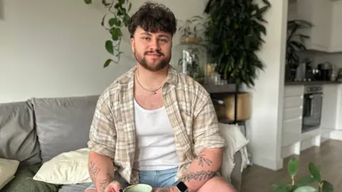 BBC A man with dark brown hair and a beard sits on a sofa smiling at the camera. He's wearing a checked beige shirt over a white vest and has tattoos