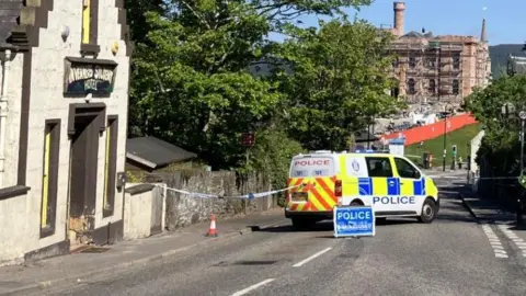 A police van is parked across the road behind blue and white police tape, and a blue sign with the word "police" on it. Inverness Castle is in the background.