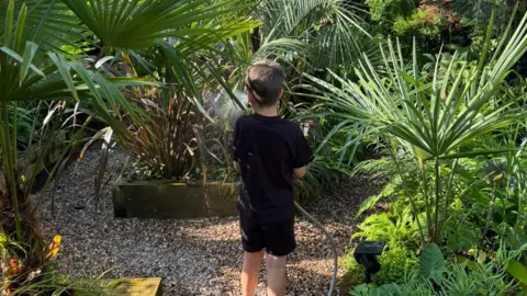 Dave Robinson Dave and Gemma's son Arlo is photographed with his back facing the camera. He is wearing a black t-shirt with matching shorts. Arlo is holding a hosepipe and is watering the plants. 