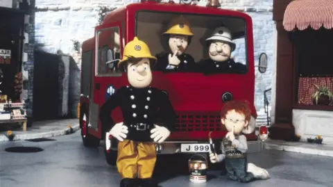 Mattel A picture from the 1987 Fireman Sam series. Fireman Sam is stood in front of the fire engine Jupiter which has the license plate J999 on the front. There are other characters sat in the vehicle and a child also in front. 