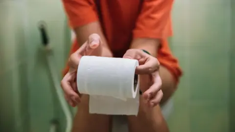 Woman sitting on toilet holding toilet paper roll - stock photo