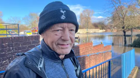 Paul Lacey standing next to the flooded river. He is wearing a Spurs beanie and a waterproof jacket.