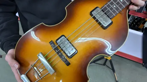 The violin-shaped body of a Hofner bass guitar being held in an unseen person's hands. It is an orange and brown colour, sometimes called 'sunburst'.