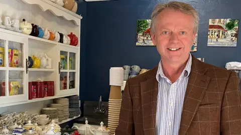 Andrew Byne smiling into the camera. He has grey, short hair and is wearing a blue and white striped shirt underneath a brown, tweed blazer. He is in front of a counter that is covered in tea pots and tea cups. There are paper coffee cups, stacks of plates and boxes of tea too. The wall behind him is navy blue which paintings of a street.