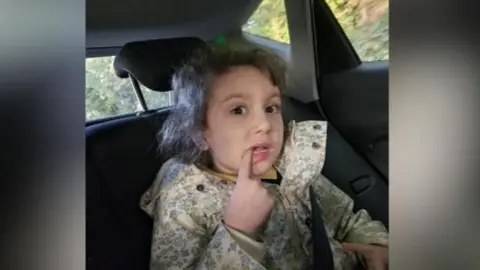 Columba Mailey Maisie is sitting in the back seat of a car. She has curly brown hair and wears a light flowered raincoat. She has the index finger of her right hand in her mouth, pointing at her tooth.