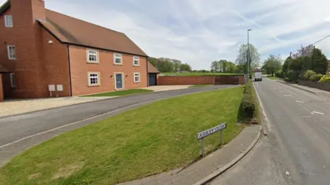 A street  view image showing a sign on the ground says "Ashley Close" with a small patch of grass behind it. Behind the grass is a driveway in front of a two-storey detached house. On the right is a main road running through a rural village.