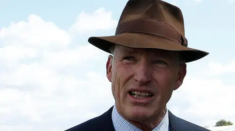 A head and shoulders image of trainer John Gosden. He is wearing a navy blazer over a shirt and a tie and a brown hat. He is standing on a racecourse. A racecourse stand - filled with people - is on the right of the image.