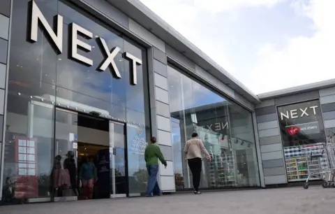 Chris Ratcliffe / Bloomberg via Getty Images A Next store is seen with the logo looming large over passers-by as they walk outside the storefront on a sunny-cloudy day in Basildon in October.
