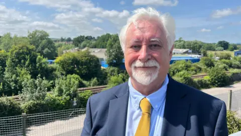 A man with grey hair and a grey goatee in a blue suit and shirt, with a golden yellow tie standing in front of a railway line.