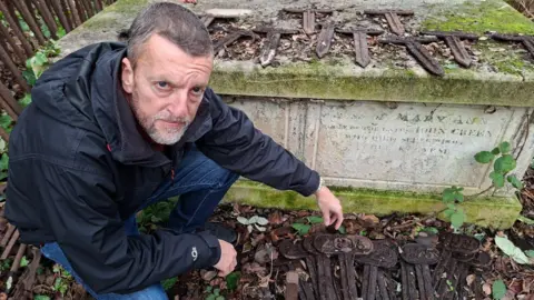 Henry Godfrey-Evans/BBC A man knelt down next to a cluster of rusted iron tags, each one has a faint number engraved on it. There is a grave, or tomb, behind him.