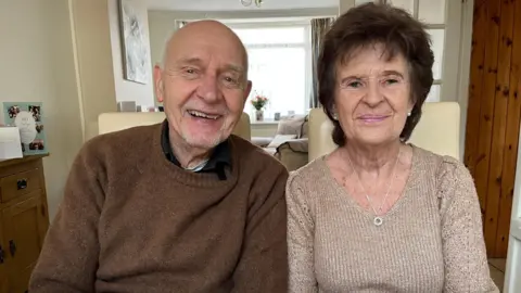 Nicola Haseler/BBC A man on the left in a brown jumper smiles at the camera. A woman sits next to him with brown hair and a beige jumper also smiling. 