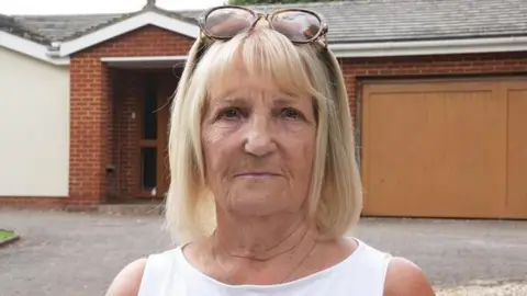 Veronica Strathie is in the middle of the image and is standing in front of a bungalow. She has shoulder length blonde hair and brown sunglasses on her head, and is wearing a white T-shirt. 