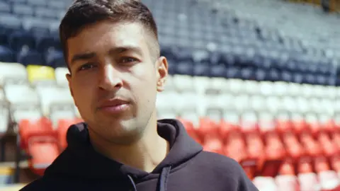 Head and shoulders image of Manpreet Singh. He has short dark hair and is wearing a navy hoodie. He's standing in front of black, red and white seats in a football stadium.