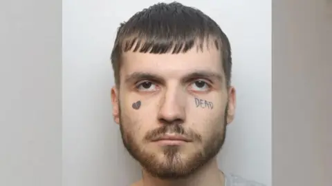 Derbyshire Police Custody image of Jack Towell. He has brown hair, brown facial hair and two facial tattoos. One is a broken love heart and the other is the word 'DEAD' under this left eye.