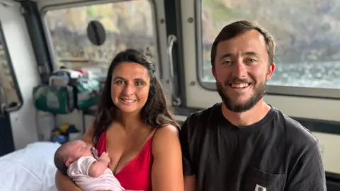 A man and woman looking at the camera and smiling. The woman is holding a new born baby in her arms.