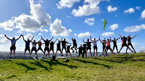 A group of runners on a hilltop, jumping up in the air with their arms out-stretched. The blue sky is dotted with clouds, there is grass in the foreground.