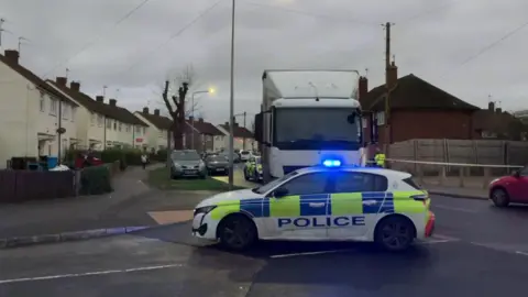 Police car with blue lights in front of a white HGV. There are houses and cars in the background. 