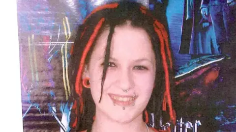 Lancashire Police/PA Wire A photograph of a young woman smiling, stood against a graffiti-style backdrop. She has red and black dreadlocks, multiple lip piercings, a nose ring and a large stretcher-style earring.