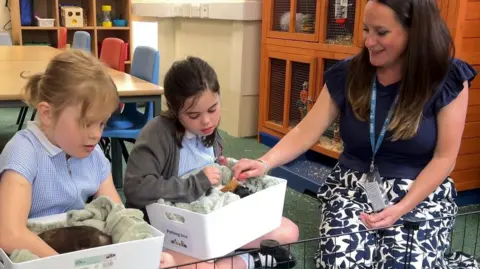 Nancy, who has blond hair in a ponytail and is wearing a blue gingham dress, sits on the left of the picture, she is holding a white box containing a black-furred guinea pig and is leaning in to stroke him. Isabelle is in the middle, wearing a grey cardigan and blue gingham dress. She has black hair which is also tied back, she is also holding a white box with a label which says "petting box" inside is a long-haired guinea pig, with black, white and brown spots. Their teacher, Caroline Sims, sits to the right of the picture, she has long brown hair and is wearing a blue top with frilled sleeves and white trousers with a blue leaf pattern. She is reaching into the box being held by Isabelle to stroke the guinea pig.