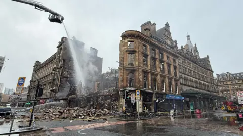 The fire-ruined blackened remains of a sandstone building on the corner of a junction in Glasgow city centre. The scene is cordoned off with yellow tape and the building's rubble surrounds the building perimeter.