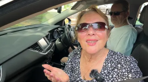 A woman is wearing sunglasses while speaking from the passenger seat of a car. She has a microphone clipped to her dress.
