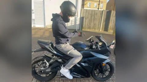 Supplied Harry Pyett sits on his motorcycle. He is wearing a helmet, hoodie, combat trousers and white trainers. His bike is a sporty-looking model in black and silver.