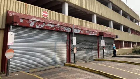 Shutters closed outside a car park, with a man looking at a sign. The building is mostly concrete.