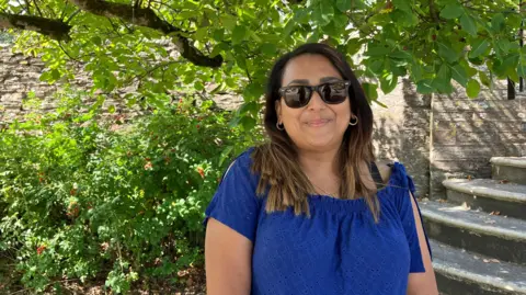 A woman, with short brown hair, wearing a blue dress and sunglasses. Behind her is steps to the right and a tree. The leaves are green. There is a brick wall behind her.