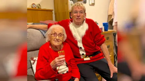 Geoff Stonebanks A man and his elderly mother. Both are wearing Christmas outfits. 