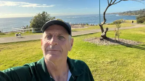 Peter Meagher / Facebook Peter Meagher wearing a green polo shirt taking a selfie in a cap by the sea
