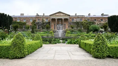 Tim Rooke/REUTERS A wide view of Hillsborough Castle and its garden courtyard on a cloudy day. The castle is a large, detached two and three-storey multi-bay neoclassical house with a red sandstone ashlar exterior. A circular fountain and a set of stone steps lead up from the garden to the castle's central portico. The landscaped courtyard contains neat box hedging and tall Irish yew trees among other plants and shrubs.