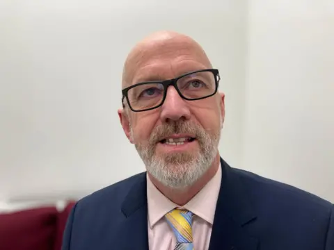 A head and shoulders photograph of council leader Craig Leyland. He has black glasses, a bald head and a grey beard. He has a pink shirt on and a multi-coloured tie under a blue suit jacket.